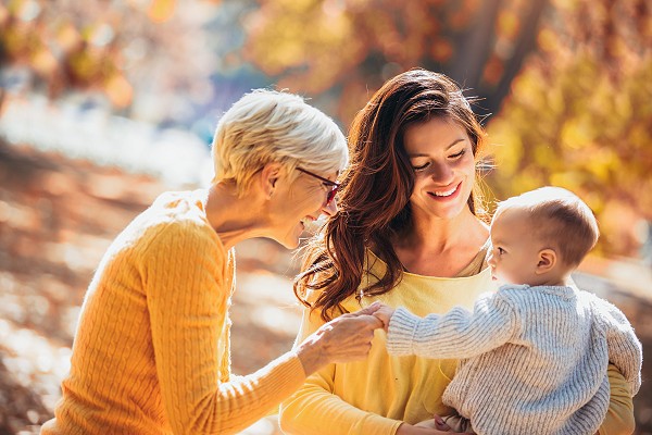 Grandmother and mother smiling at baby in autumn park.